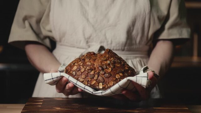 Close-up of baker in a rustic linen apron holding freshly baked artisan bread topped with pumpkin seeds on a checkered towel. Homemade loaf for bakery promos, organic product and healthy food concept.