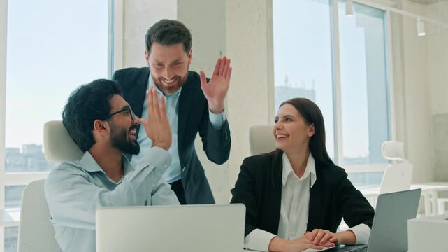 Happy multiethnic business team working laptops indoor office leader shows papers and thumb up celebrating deal success give high five teamwork excited diverse coworkers men woman computer achievement