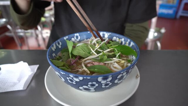 Close up shot of person using chopsticks to stir noodle soup in restaurant setting