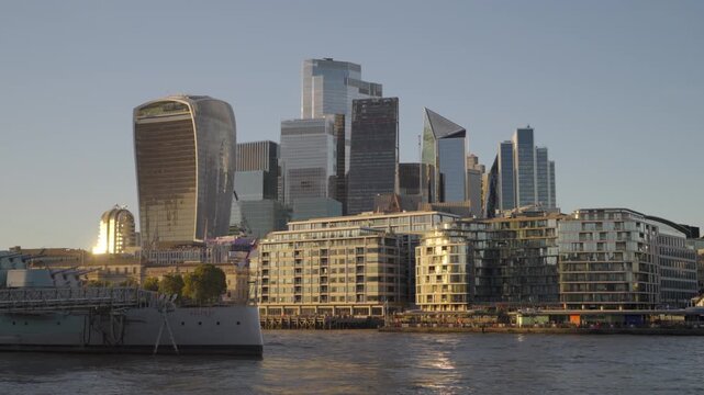 Low-angle close-up view across the River Thames framing the modern skyscrapers of the City of London financial district and HMS Belfast in late afternoon sun.