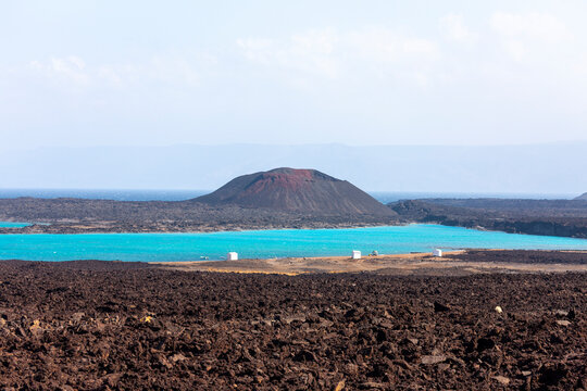 A red-tinged volcanic cone overlooks a turquoise lagoon bordered by black lava fields, Tadjourah Region, Goubet Al-kharab, Djibouti