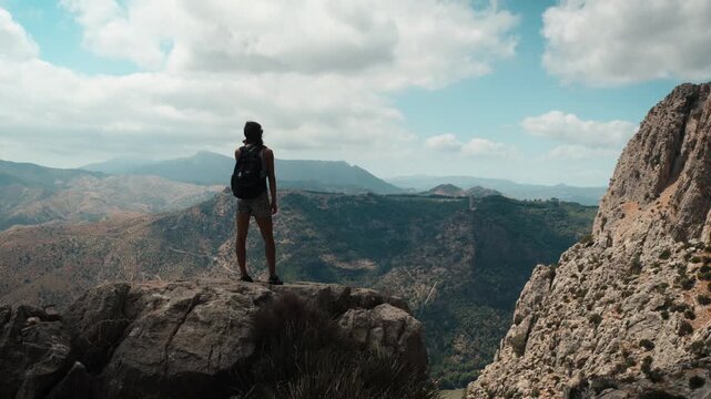 A young woman with a backpack standing on a rocky cliff edge facing expansive mountain ridges and a deep valley under a cloudy sky in El Chorro Andalusia Spain