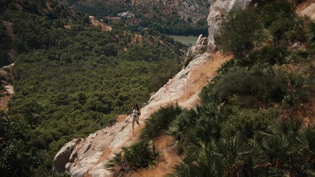 A young woman hiking along a steep narrow dirt path on a rocky mountainside surrounded by dense forest and distant hills in Andalusia Spain under daylight on a solo mountain trek