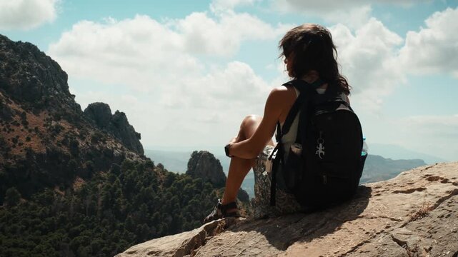 A woman with a backpack sitting on a sunlit rocky ledge gazing over a pine covered valley and distant limestone cliffs under a partly cloudy sky in El Chorro Andalusia Spain