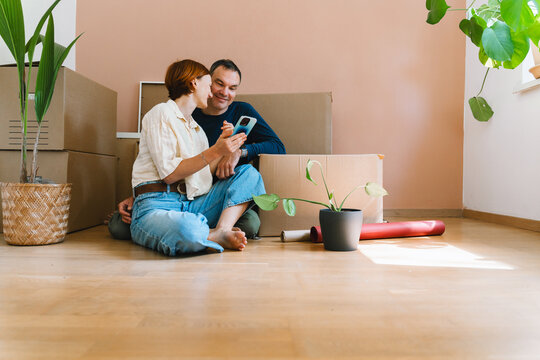 A mature caucasian couple looks at a smartphone screen together while sitting on the floor by moving boxes. Digital housing search and modern family lifestyle concept.