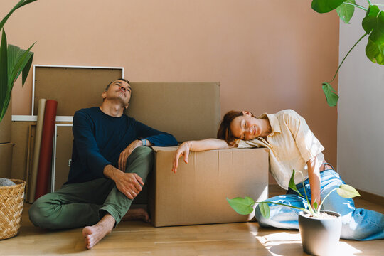 A mature caucasian couple rests exhaustedly against cardboard boxes during their busy moving day. Relocation fatigue and realistic domestic lifestyle concept.
