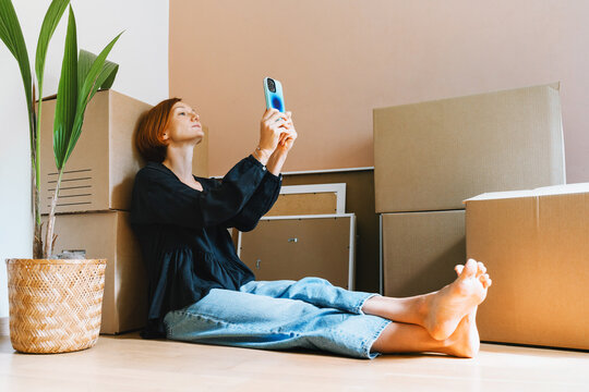 A mature caucasian woman sits on the floor among cardboard boxes using her smartphone device. Easy relocation process and modern digital lifestyle concept.
