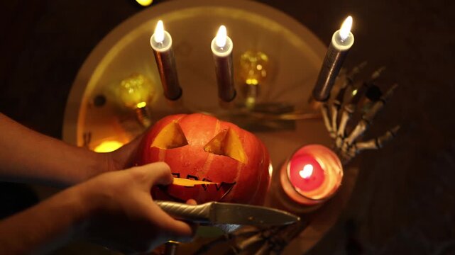 Halloween pumpkin carving continues as hands scoop pulp from a smiling jack o lantern surrounded by candles and skeleton props during a creative October party preparation
