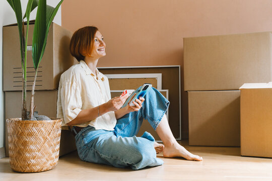 A mature caucasian woman holds a credit card and smartphone while sitting by unpacked moving boxes. Online shopping and remote household purchasing concept.