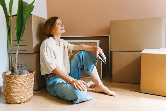 A mature caucasian woman sits on the floor among cardboard boxes with smartphone device in hand. Easy relocation process and modern digital lifestyle concept.