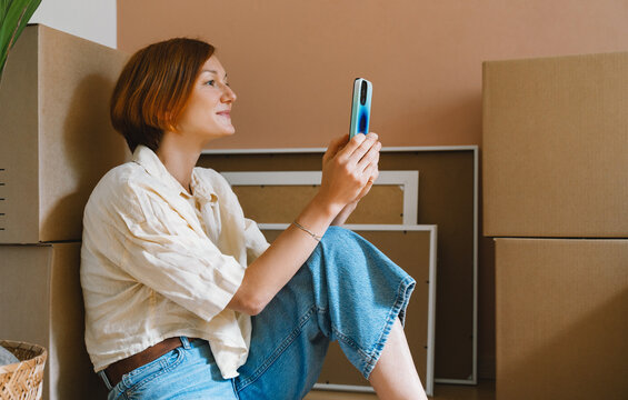A mature caucasian woman sits on the floor among cardboard boxes using her smartphone device. Easy relocation process and modern digital lifestyle concept.