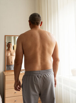 An overweight, middle-aged man stands shirtless with his back to camera, looking at his reflection in mirror and assessing changes in his physique. Real person, self-acceptance, authentic lifestyle