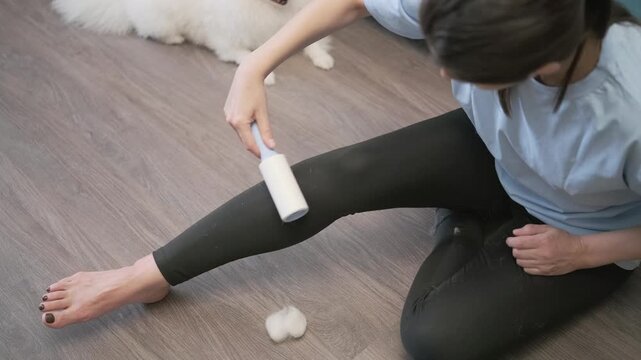 Seated woman carefully cleans black leggings with lint roller, moving handheld tool along thigh and shin while removing cotton tufts and pet fur during indoor clothing maintenance routine