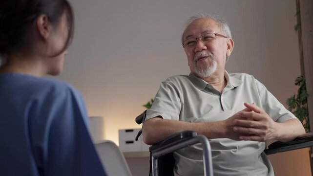 Asian elderly man in a wheelchair smiles and talks with caregiver during a friendly home visit, sharing a warm and trusting conversation together indoors.