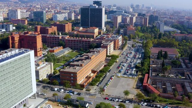 Aerial drone view of red brick university campus buildings in Zona Universitaria, Les Corts district, with tree-lined avenue, parked cars and city skyline, Barcelona, Spain, on a sunny day.
