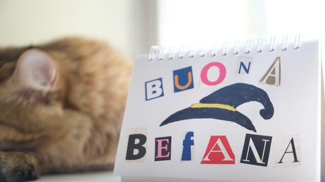 Cozy home moment: cute cat napping beside a child&rsquo;s handmade &ldquo;Buona Befana&rdquo; greeting. The letters are cut from magazines and carefully glued onto the paper, giving the note a tender, handmade charm.