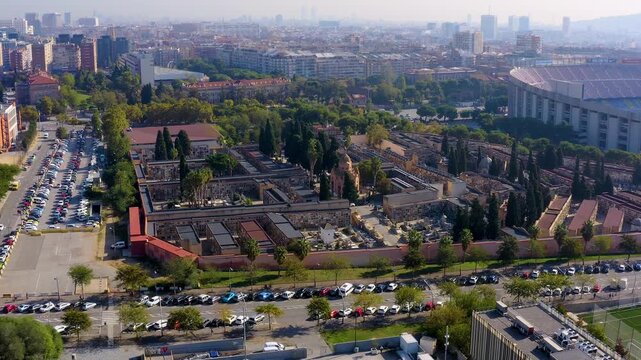 Aerial drone view of historic Les Corts Cemetery with cypress trees and niche walls beside the old Camp Nou stadium, city skyline in haze, Barcelona, Spain, on a sunny day.