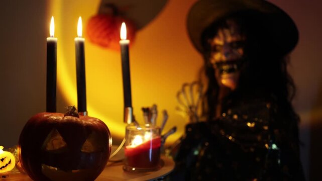 Halloween woman in skeleton makeup and witch costume scares with skeletal hands beside carved pumpkin and burning candles creating a creepy October party scene indoors at night