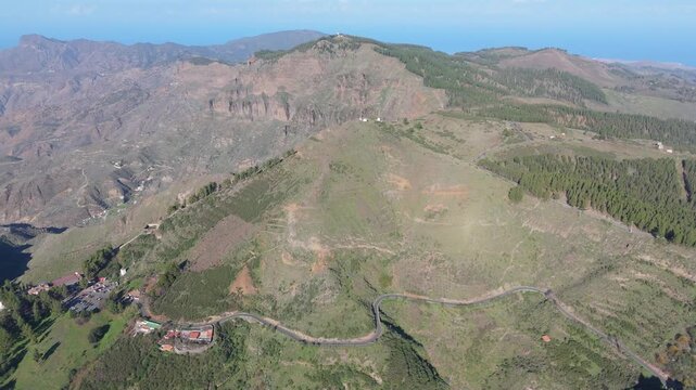 An aerial drone view pans right over the rugged mountainous landscape of Gran Canaria. The shot features the hamlet of Cruz de Tejeda, winding roads, lush green hills, and the distant ocean.