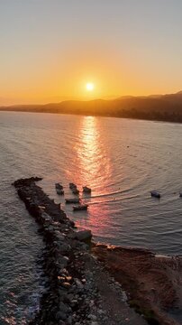 Aerial view of stone breakwater jetty and moored boats in calm sea water during golden hour sunset at Roda beach in North Corfu, Greece vertical video