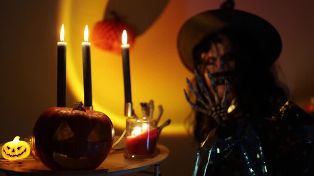 Halloween woman in skeleton makeup and witch costume poses with a skeletal hand beside carved pumpkin and glowing candles creating spooky October party atmosphere indoors