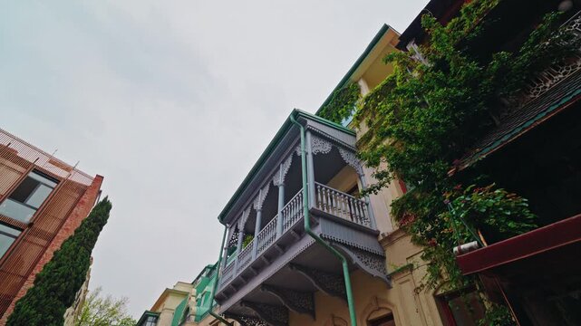 A dark carved wooden balcony with white fretwork stands under climbing ivy on the facade of an old Tbilisi house.