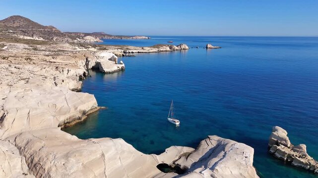 Sarakiniko displays smooth white volcanic tuff, eroded caves and sculpted shoreline on Milos, Cyclades, Greece, with an anchored yacht offshore, drone forward flyover