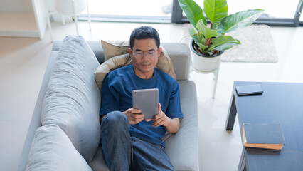 A man in a blue shirt relaxing on a grey sofa while using a digital tablet in his living room © Naypong Studio