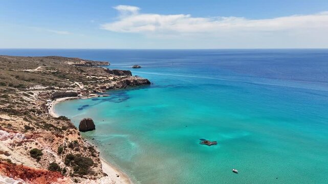 Firiplaka Beach curves along the volcanic south coast of Milos with turquoise shallows, pale rocky terrain and small offshore rocks facing the Aegean Sea, drone forward flyover