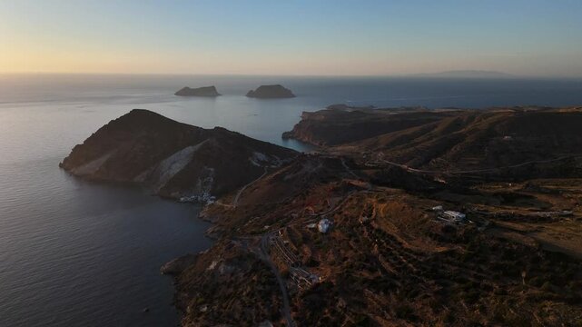 Plaka overlooks rugged volcanic slopes, the Milos caldera coastline and offshore islets near Adamas beneath the setting sun, with Antimilos visible toward the western horizon, drone flyover, Greece