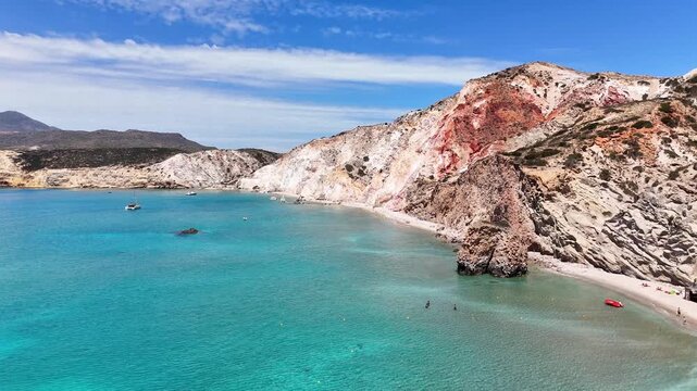 Firiplaka Beach lies beneath red, ochre and white volcanic cliffs in Milos Municipality, Milos, South Aegean, Greece, with swimmers along the sheltered bay, drone slide