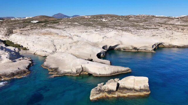 Sarakiniko exposes thick white pumice tuff and diatomite layers shaped into smooth coastal cliffs and sea caves on Milos, South Aegean, Greece, drone forward flyover