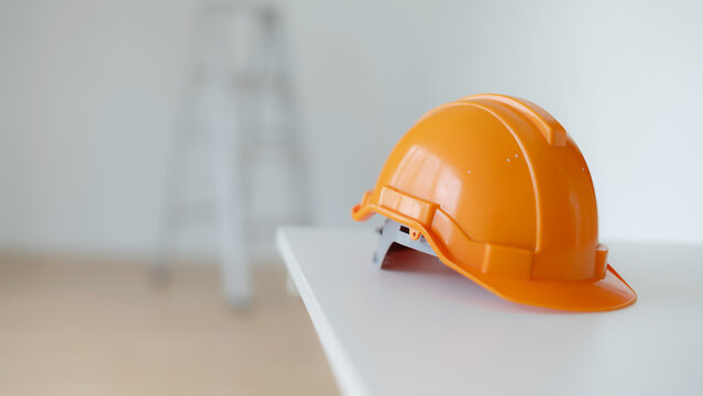 Bright orange hard hat resting on a white table with a blurred step ladder in the background