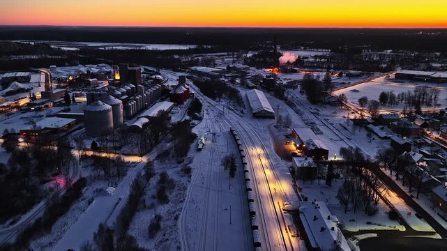 Drone flying over a snow-covered town with an industrial factory and railway at dusk