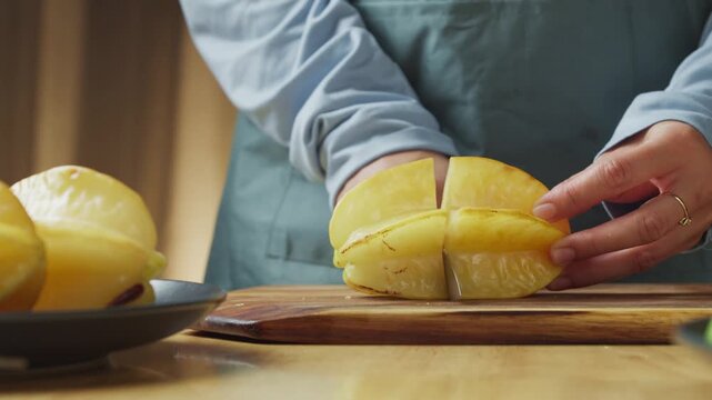 person cutting a fresh ripe yellow starfruit, also known as carambola, with a kitchen knife on a wooden cutting board