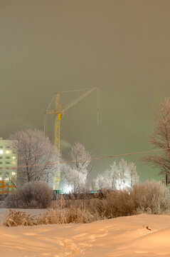 Winter urban background. A construction crane above a snow covered field. Frosted trees, a lit apartment building and glowing street lights at night