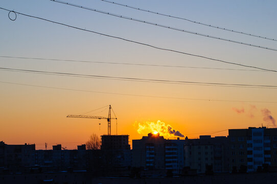 Urban sunset background. A bright sun setting behind residential buildings and a construction crane. Dark silhouettes of rooftops and steam under an orange gradient sky