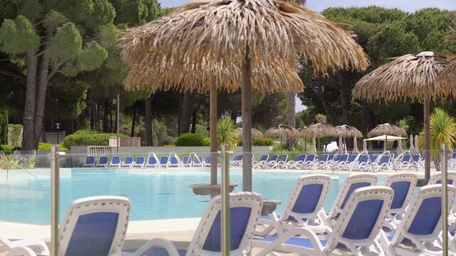 Thatched parasols and rows of blue sun loungers surround a large outdoor swimming pool