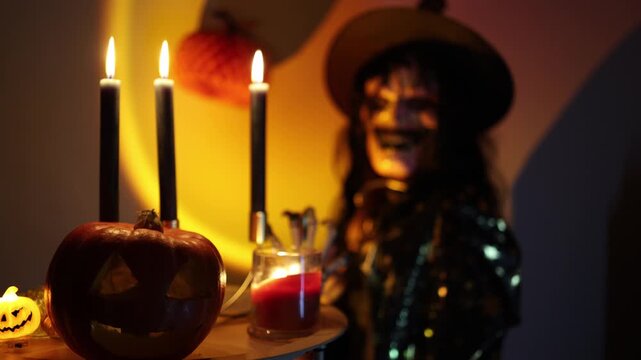 Halloween carved pumpkin and glowing candles frame a woman in skeleton face makeup and black costume creating a mysterious October party vibes in warm candlelight indoors