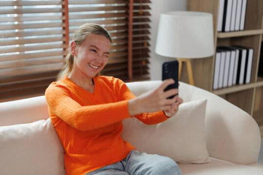 Young woman enjoying personal moments, winking and laughing while taking a selfie with a smartphone, sharing fun and authentic happiness in a cozy home environment