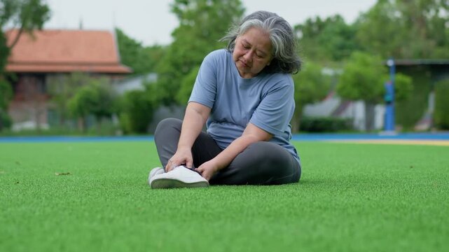 Older woman sits on a sports field holding her foot with a strained expression, illustrating foot discomfort, body awareness, senior wellness and outdoor health concerns.