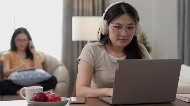 A young woman works on a laptop at home while another woman relaxes in the background. The scene represents remote work, home productivity, modern lifestyle, and work-life balance.