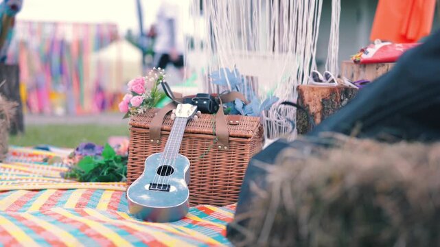 Colorful picnic scene with a silver ukulele, floral arrangement, and wicker basket on a vibrant patterned blanket at a lively outdoor festival setting