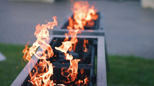 Flames dance above charcoal in a long metal grill, showcasing the vibrant fire's movement and warmth in an outdoor setting, evoking a Woodstock style atmosphere