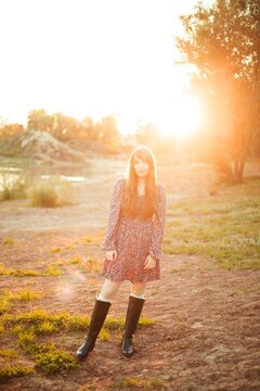Woman stands on the ground next to a river. The sun sets behind trees, creating a warm light. The person wears a dress and boots