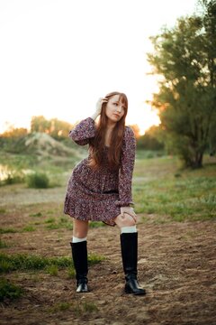 Woman stands on the ground next to a river. The sun sets behind trees, creating a warm light. The person wears a dress and boots