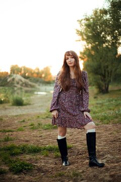 Woman stands on the ground next to a river. The sun sets behind trees, creating a warm light. The person wears a dress and boots