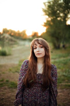 Woman stands on the ground next to a river. The sun sets behind trees, creating a warm light. The person wears a dress and boots