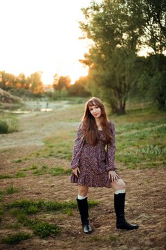 Woman stands on the ground next to a river. The sun sets behind trees, creating a warm light. The person wears a dress and boots