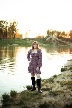 Woman standing by the water during sunset with green trees in the background